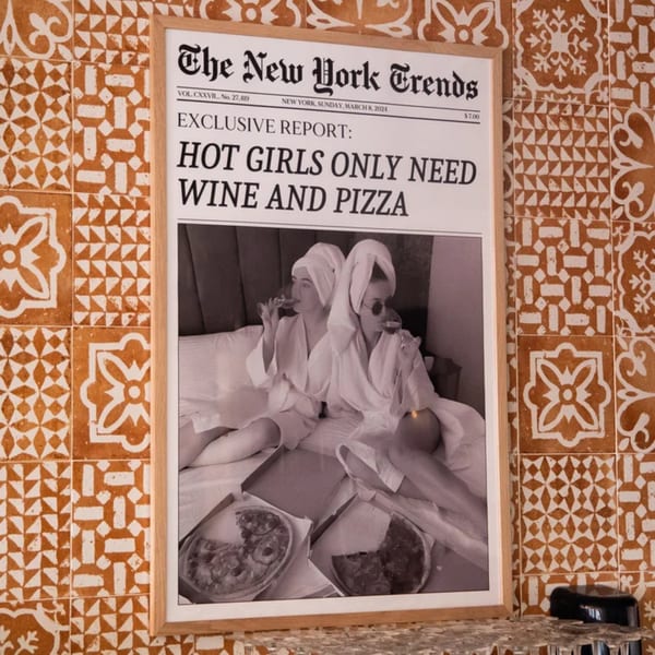 Framed newspaper article on a tiled wall, featuring two women in robes drinking wine with pizza.
