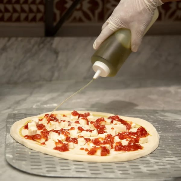 A gloved hand drizzles olive oil onto a Rocky's Pizza Bar pizza with tomato sauce and cheese.